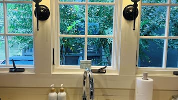 A kitchen with a stainless steel sink, two wall-mounted lamps, and a window with a view of greenery.