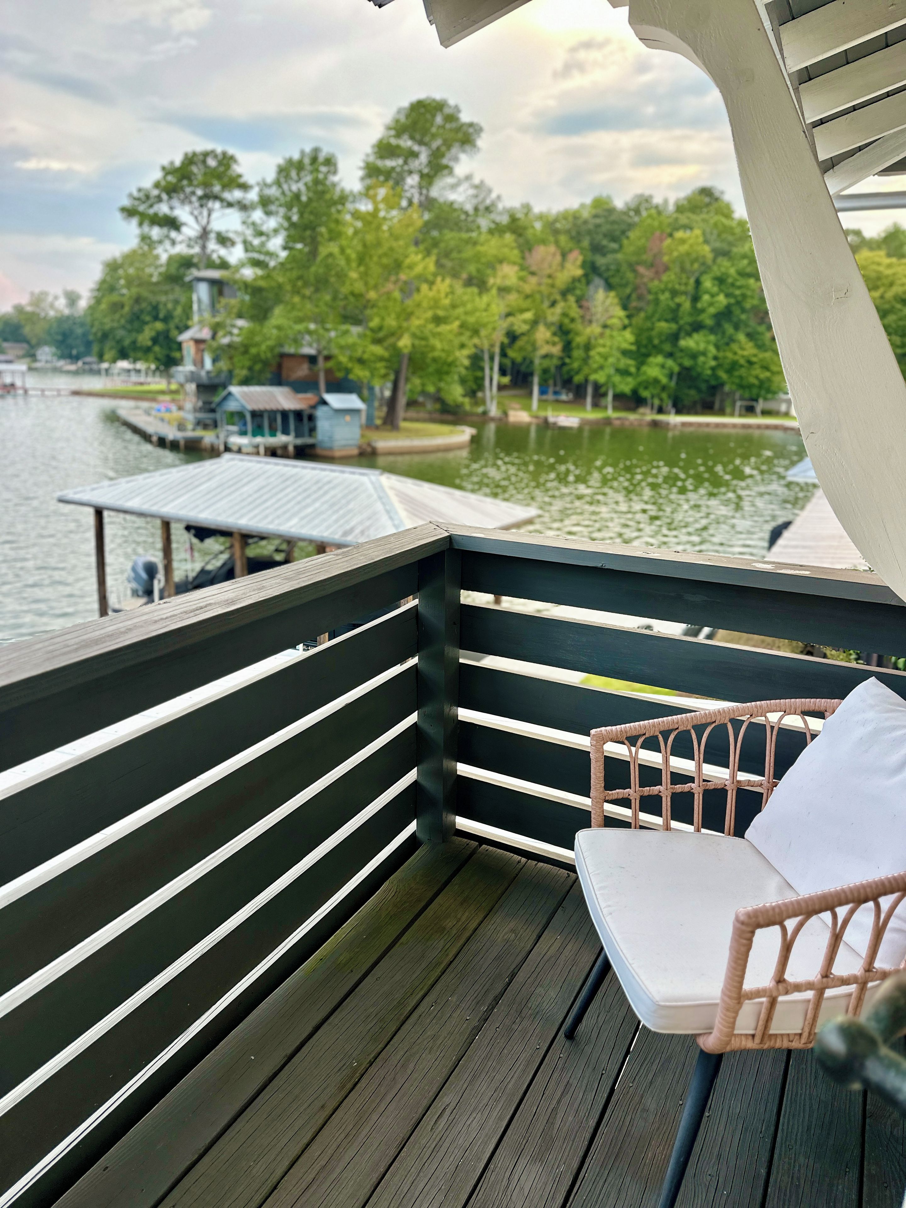 A wooden deck with a white cushioned chair, overlooking a calm body of water with docked boats and greenery in the background.