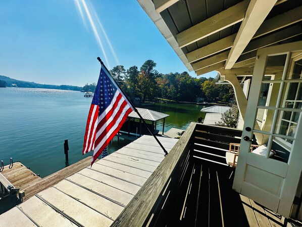 A wooden dock extending over a calm lake with an American flag, a covered seating area, and a view of a docked boat.
