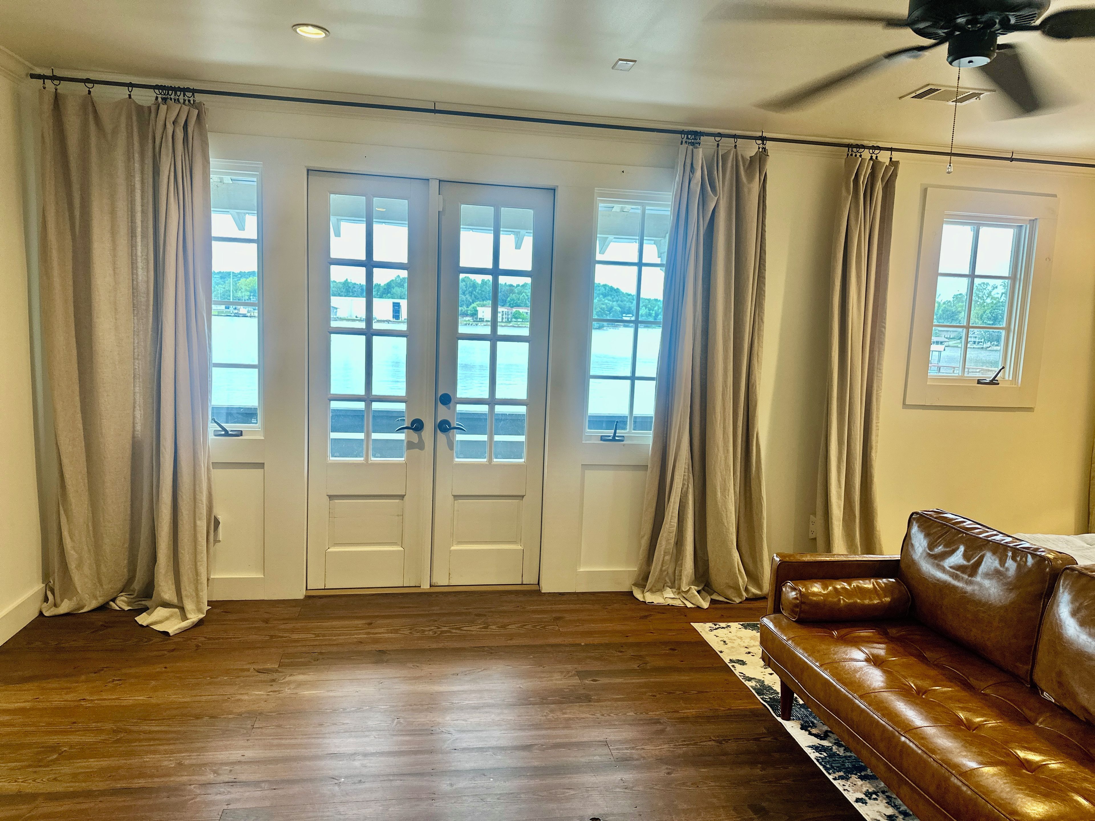 Ein Zimmer mit einem braunen Ledersofa, Holzboden, Deckenventilator und Blick auf ein Gewässer durch eine Doppeltür.