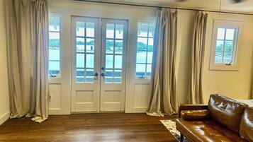 Ein Zimmer mit einem braunen Ledersofa, Holzboden, Deckenventilator und Blick auf ein Gewässer durch eine Doppeltür.