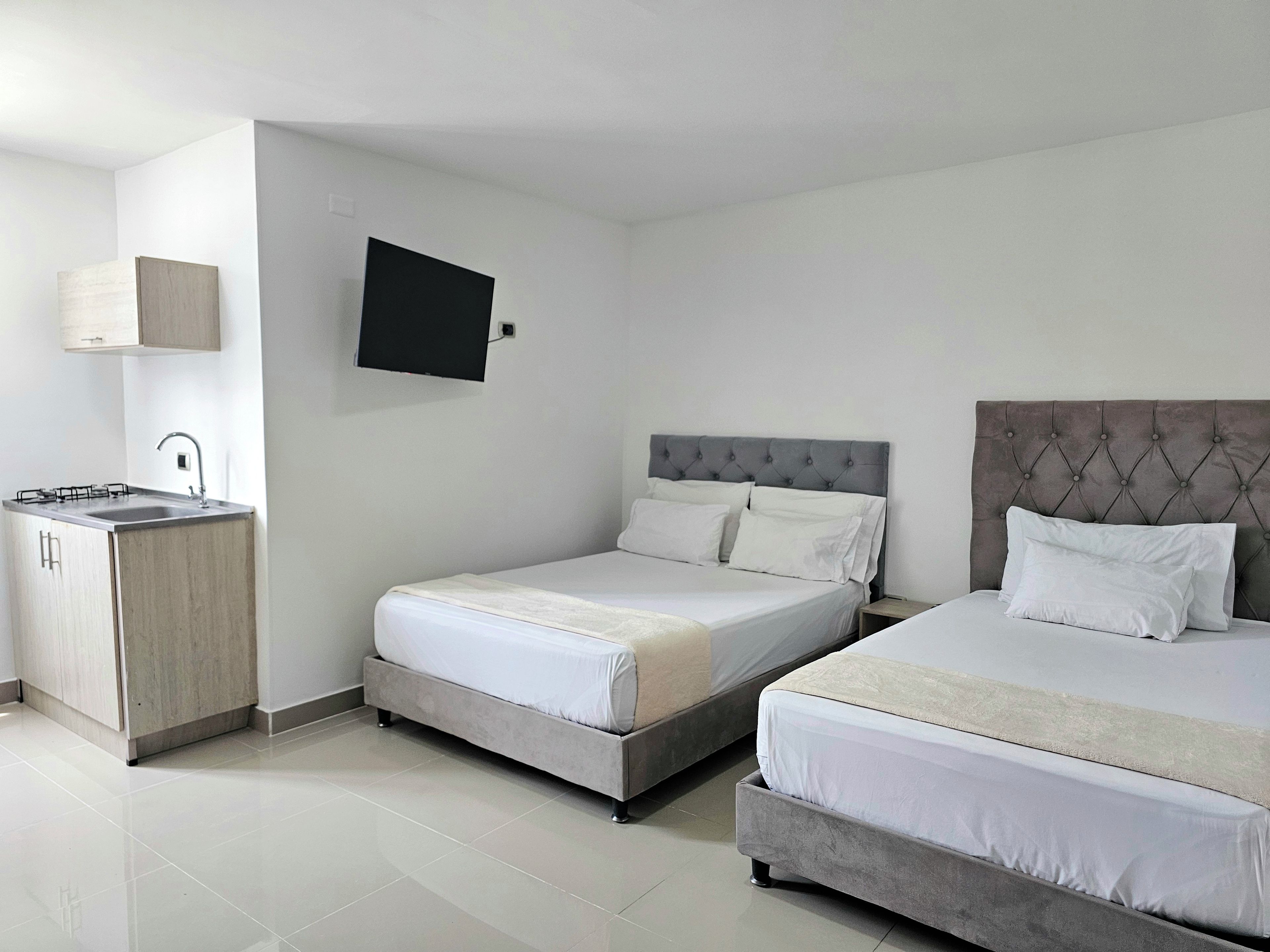 A hotel room with a kitchenette, a bed with a tufted headboard, and a flat-screen TV mounted on the wall.