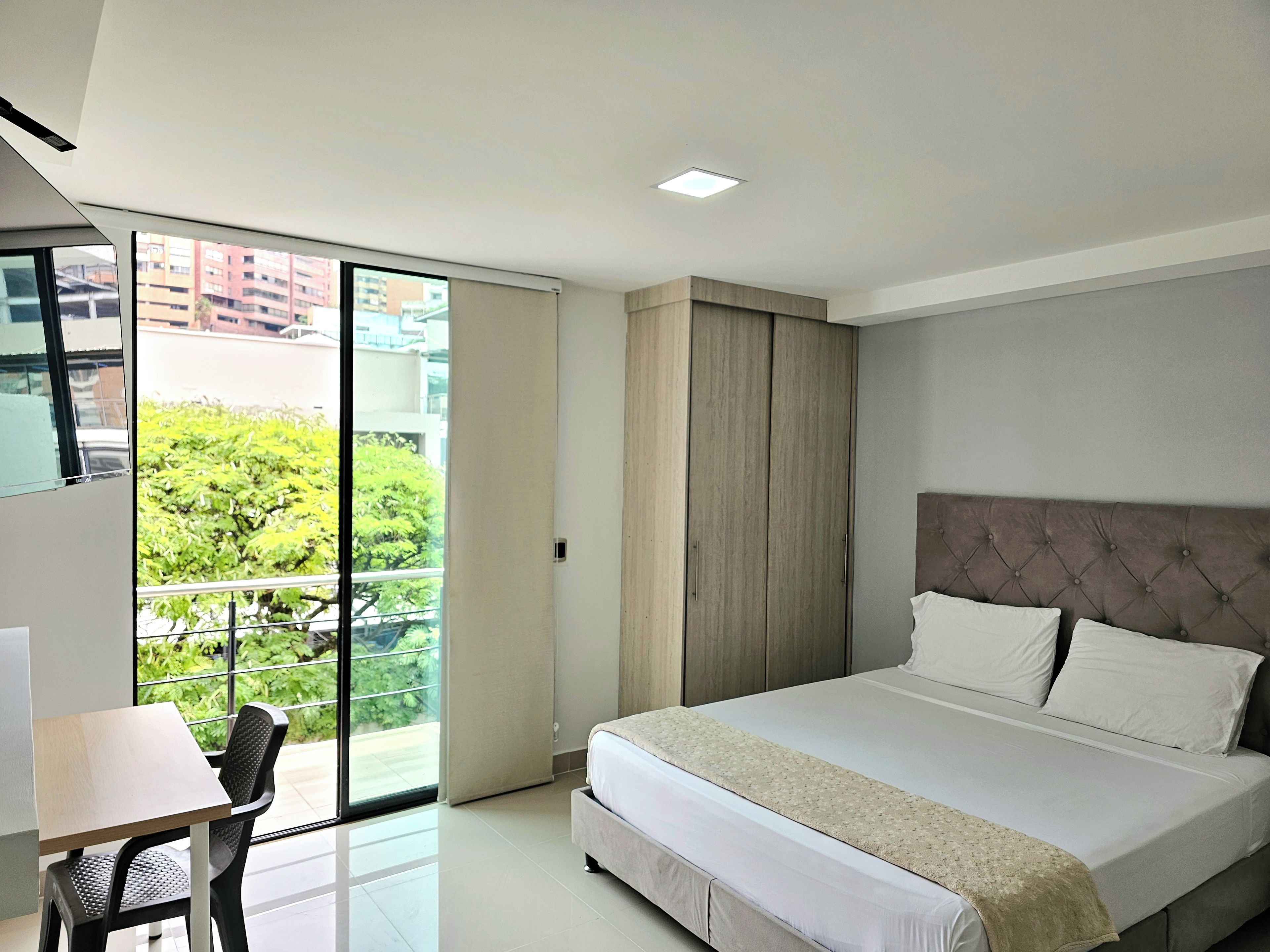 A hotel room with a bed, a desk, and a view of greenery and buildings through a large window.