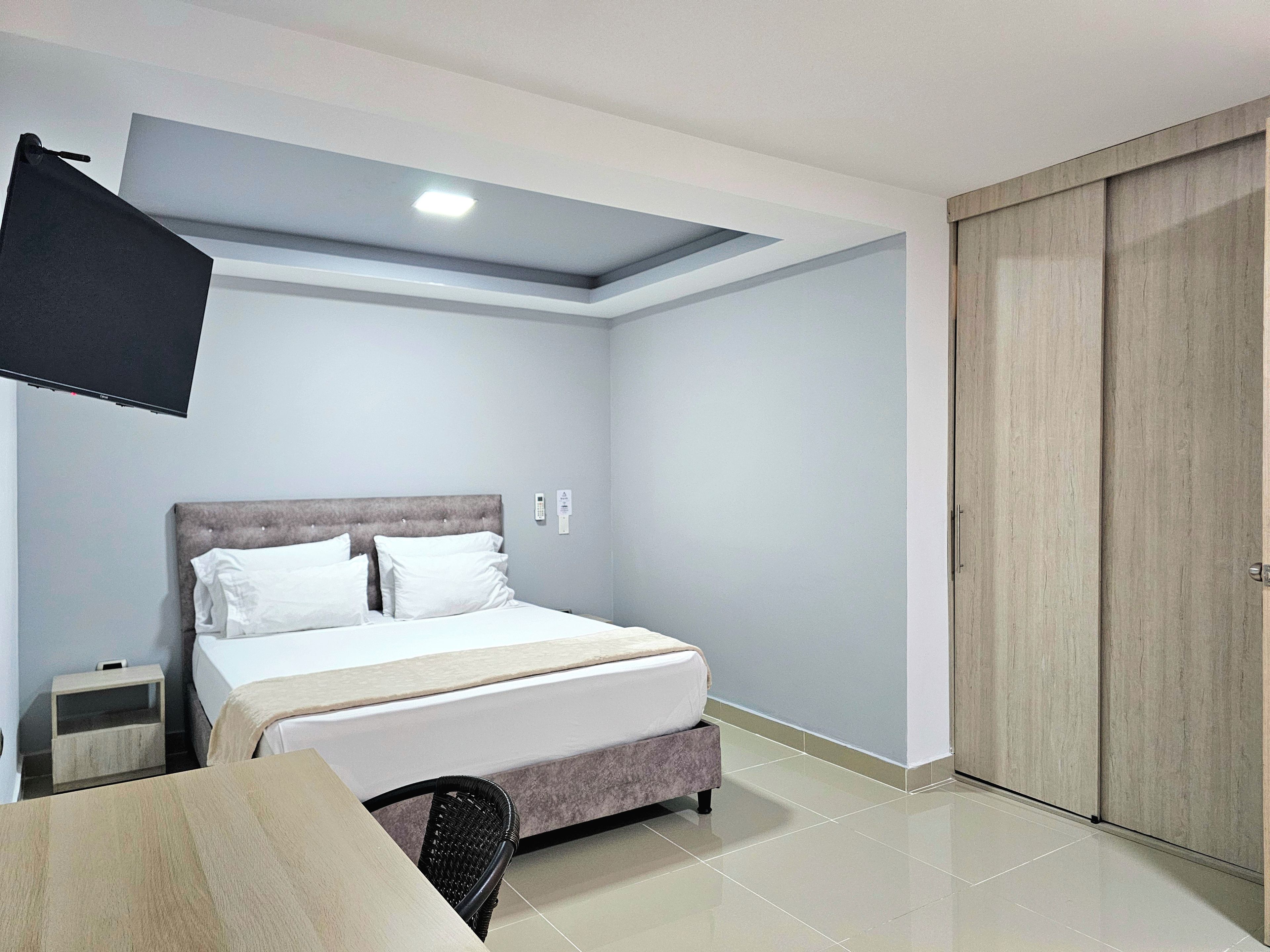 A bedroom with a bed, a television mounted on the wall, a bedside table, a wardrobe, and a chair.
