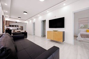 Ein modernes Wohnzimmer mit einer schwarzen Ledersofa, einem Holzschrank mit einem Flachbildfernseher und einer Küche im Hintergrund.