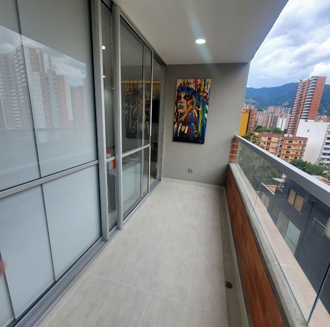Un balcón con puerta corrediza de vidrio que da a un espacio interior, un cuadro en la pared y vistas de edificios circundantes.