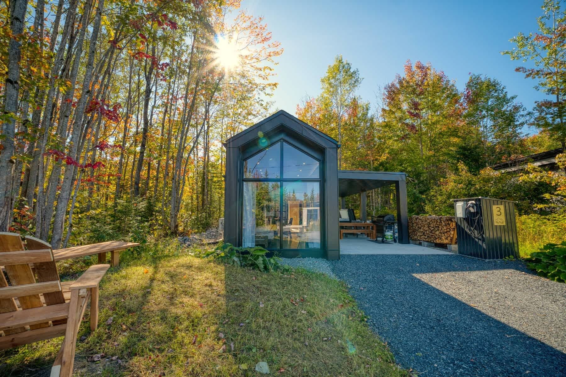 A modern cabin with a glass facade, surrounded by a forest with autumn foliage.