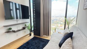A modern living room with a large flat-screen TV, a sofa, a dark blue rug, and a city view through large windows.