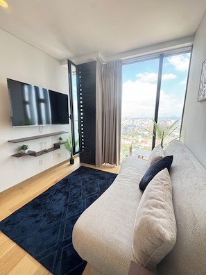 Uma sala de estar moderna com uma grande TV de tela plana, um sofá, um tapete azul escuro e vista para a cidade através de amplas janelas.