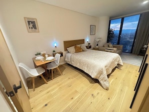Un dormitorio con cama, mesitas de noche, un escritorio con silla y un amplio ventanal con vista a la ciudad.