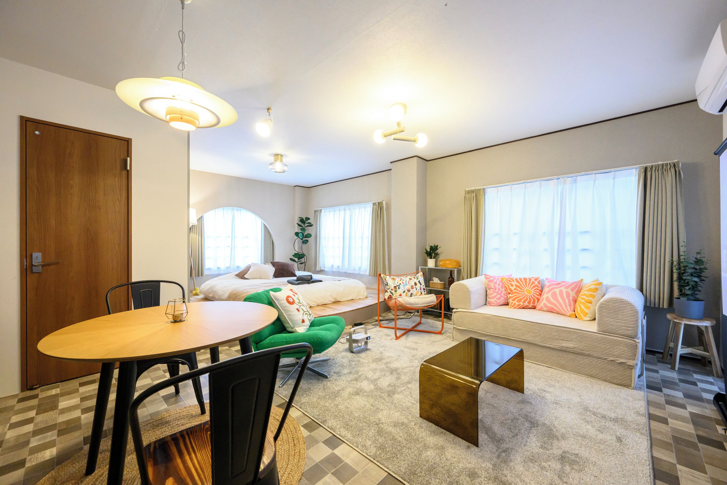 A modern hotel room with a bed, a sofa, a dining table, and a small kitchen area.