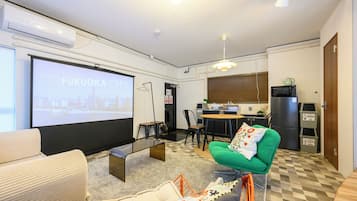 A modern living room with a projector screen displaying 'FUKUOKA CITY', a compact kitchen area, and a dining space.