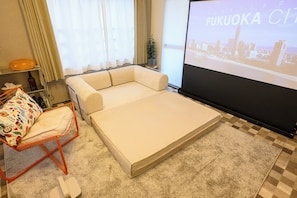 Quarto de hotel com cama, uma mesinha, uma cadeira e tela de projeção exibindo paisagem urbana.