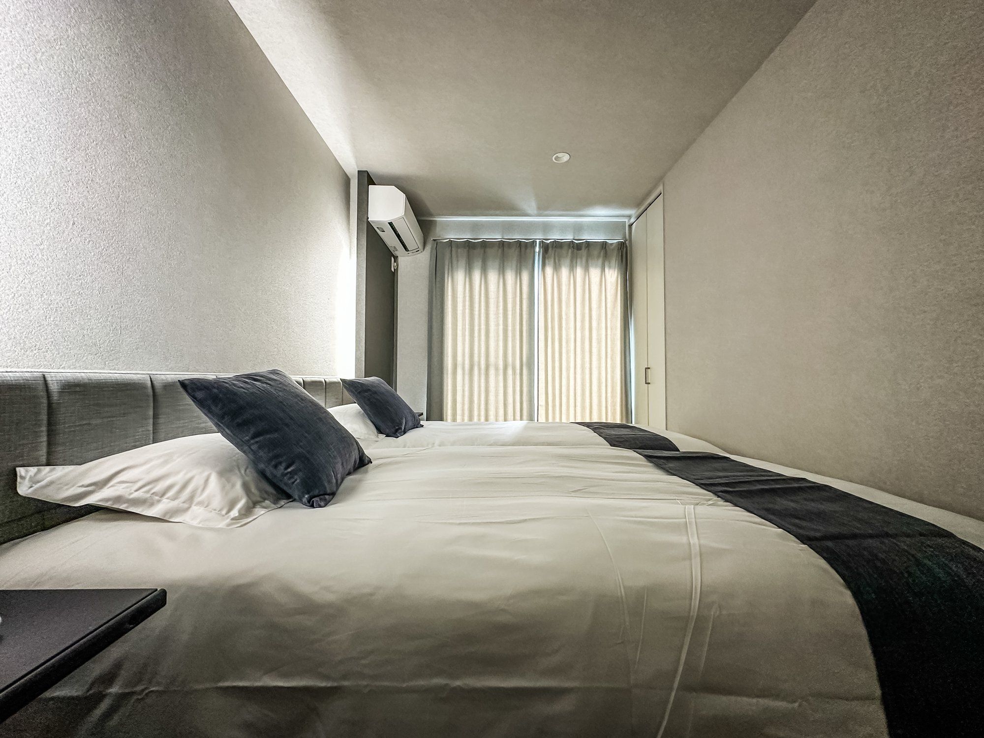 A hotel room with a bed, pillows, a bedside table, a window with curtains, and an air conditioning unit.