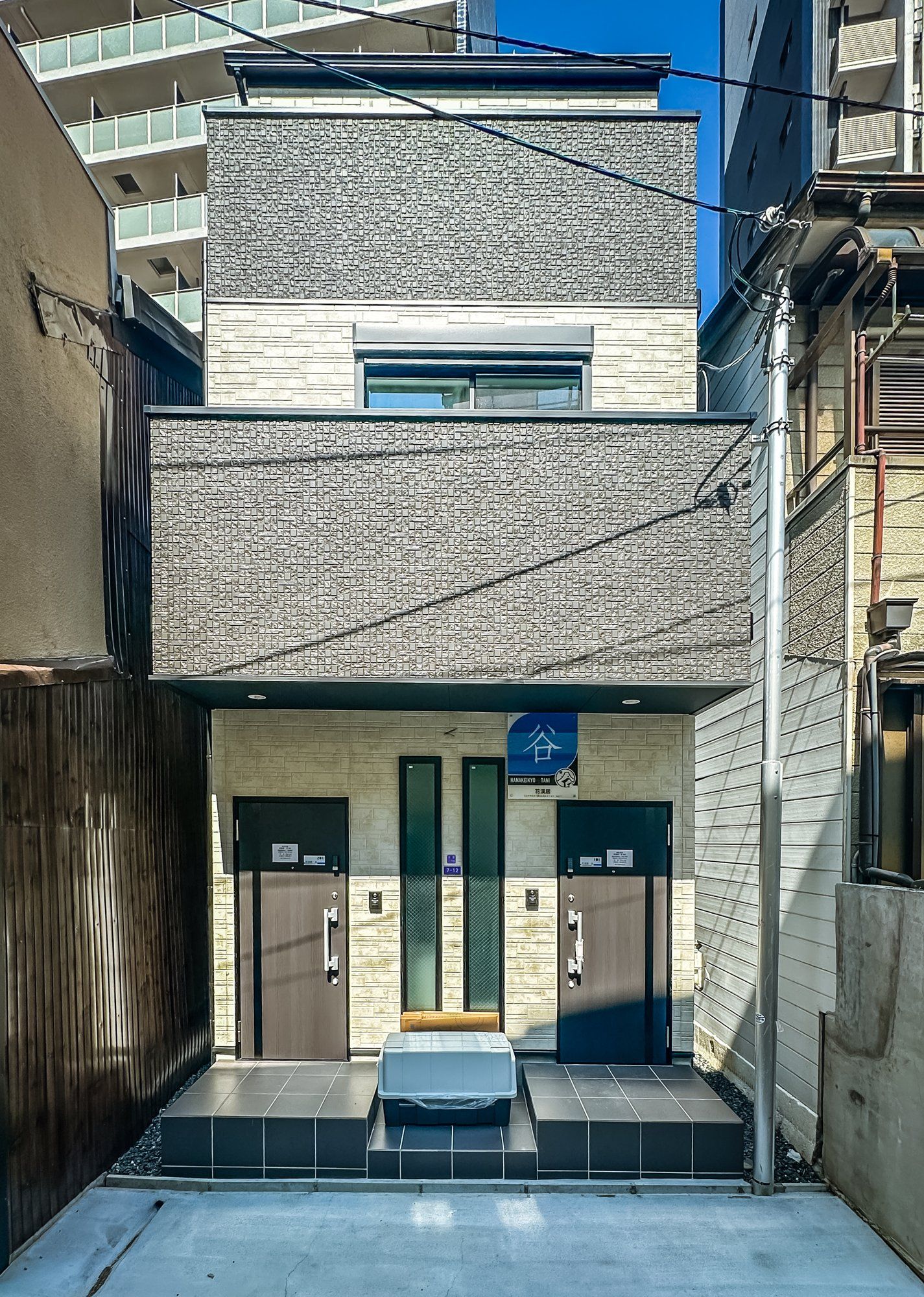 A modern building with a two-story facade, a small entrance with a blue sign, and a set of double doors.
