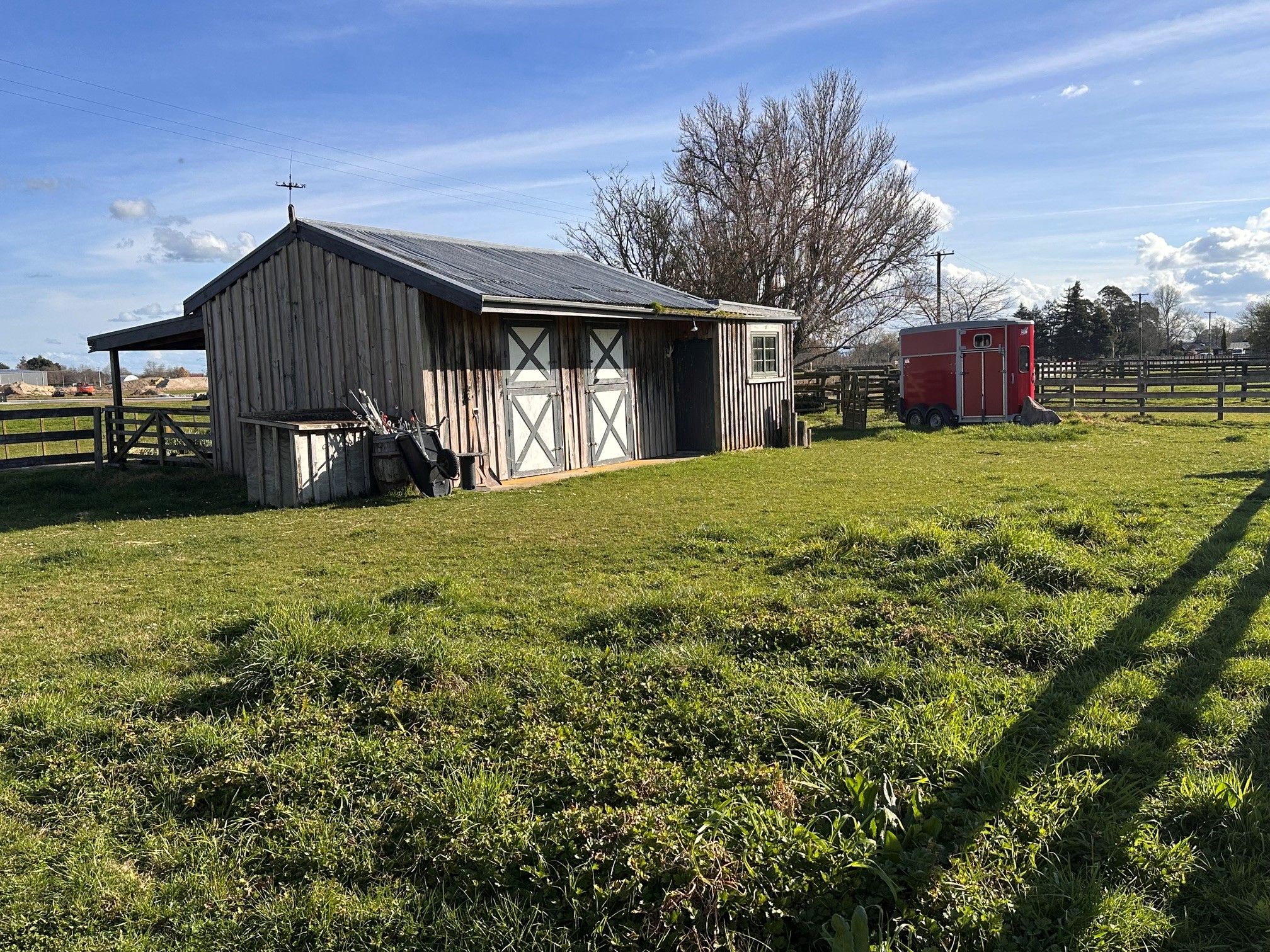 A barn with a sloped roof, a fenced area, and a red trailer in a grassy field.