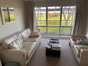 A living room with a beige sofa, a coffee table with books, and a view of a green field through large windows. - Country home - amazing wide north facing horse stud and country views (Pukemoremore)