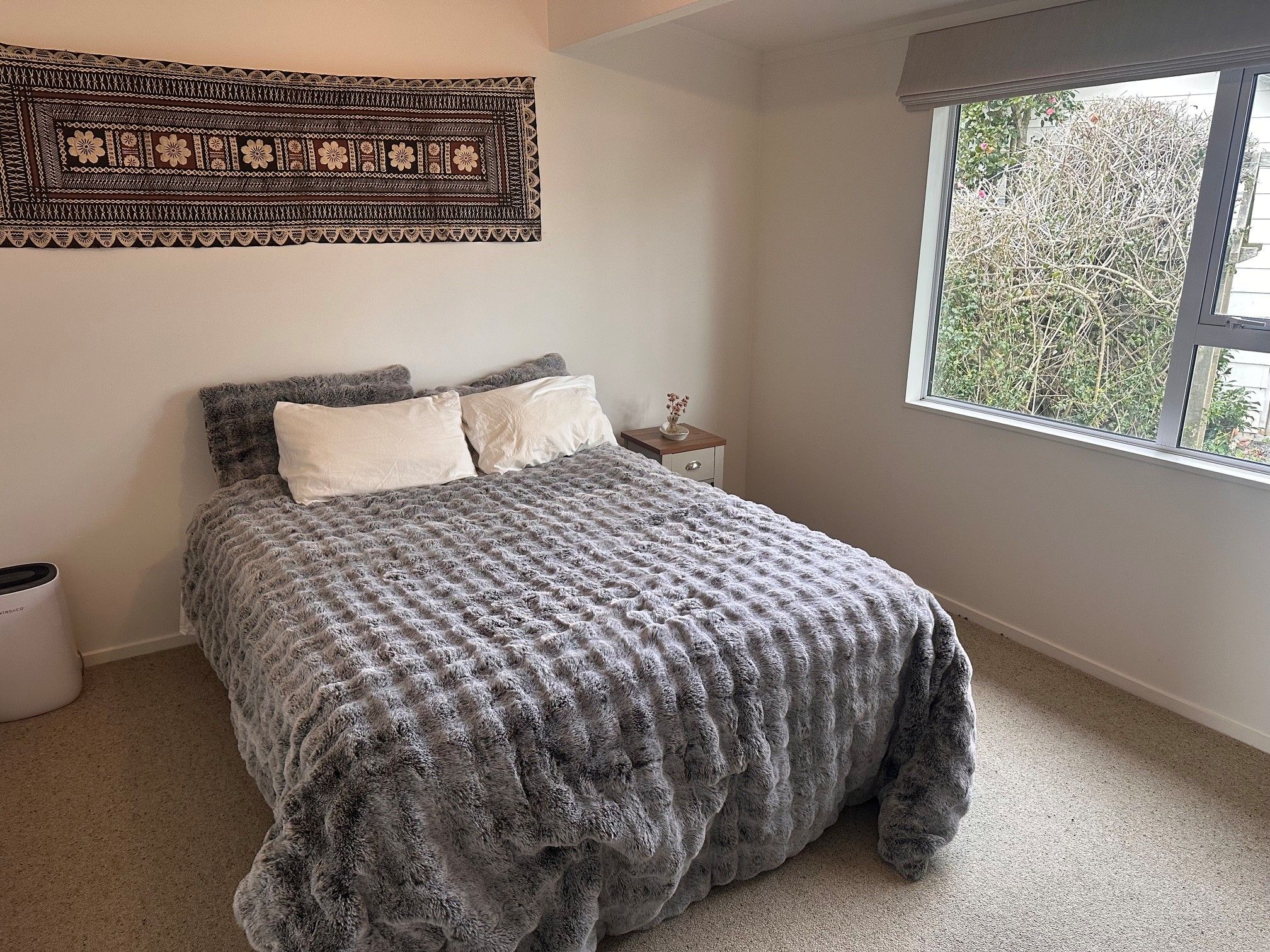 A bedroom with a bed, a nightstand, a window, and a wall hanging.
