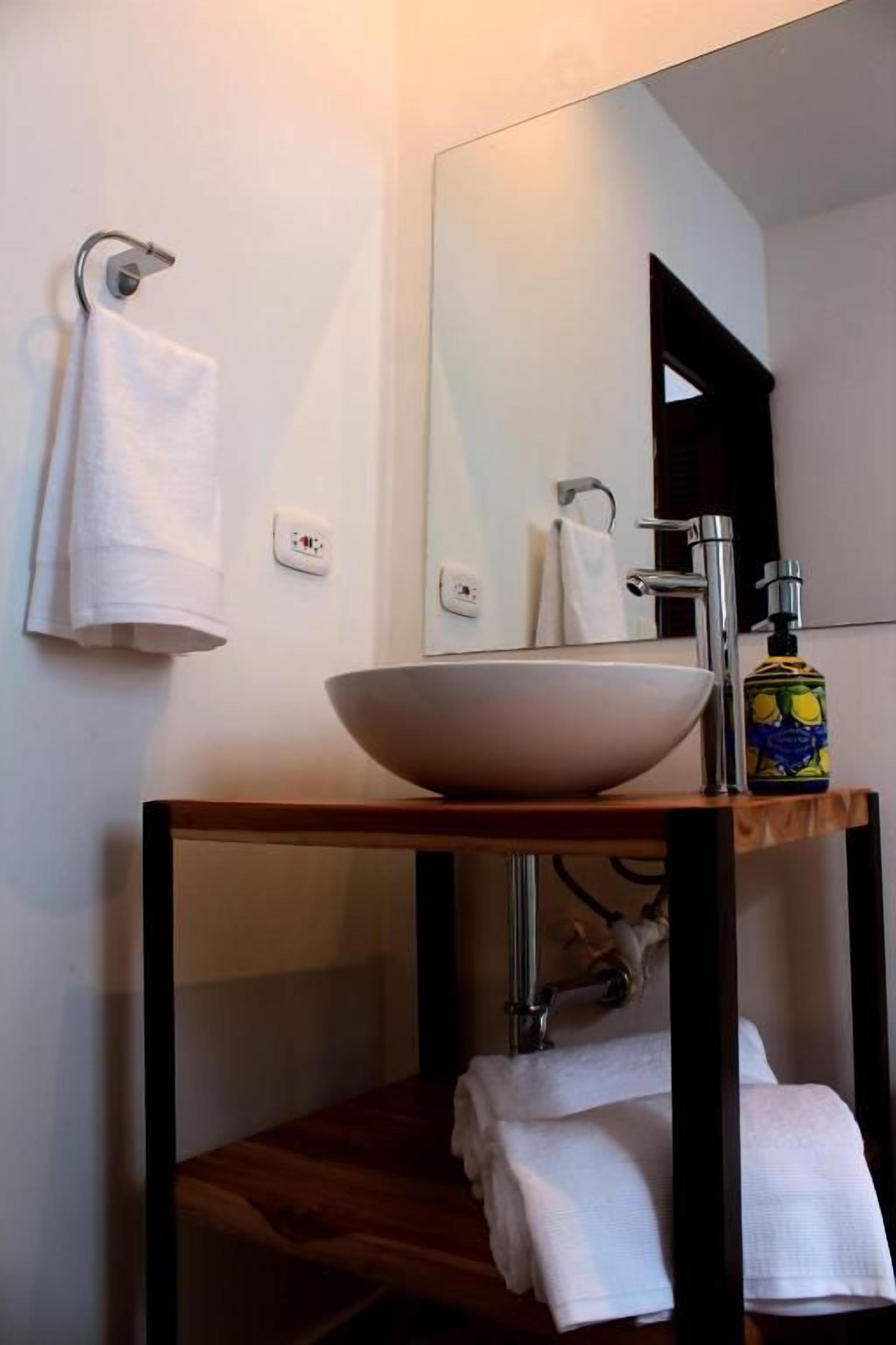 Un baño con un lavamanos blanco, un espejo, un perchero para toallas y una botella de jabón.