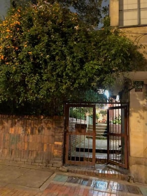 A gated entrance with a metal gate, a brick wall, and a tree with orange flowers. - Casa Muntu (Bogotá)