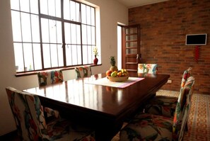 A dining area with a large wooden table, floral-patterned chairs, a fruit bowl, and a brick wall. - Casa Muntu (Bogotá)