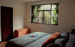 A bedroom with a bed, a window with bars, a bench, and a door. - Casa Muntu (Bogotá)