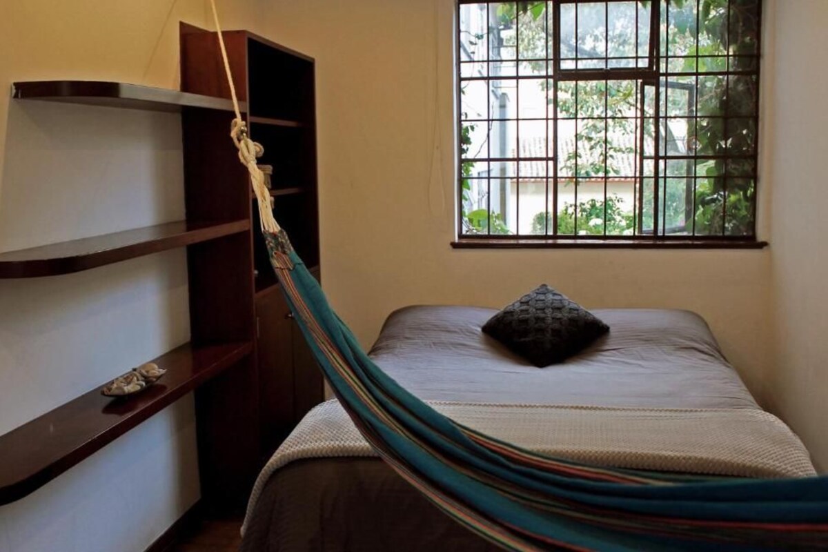 A room with a bed, a hammock, a window with a view, and a shelf.
