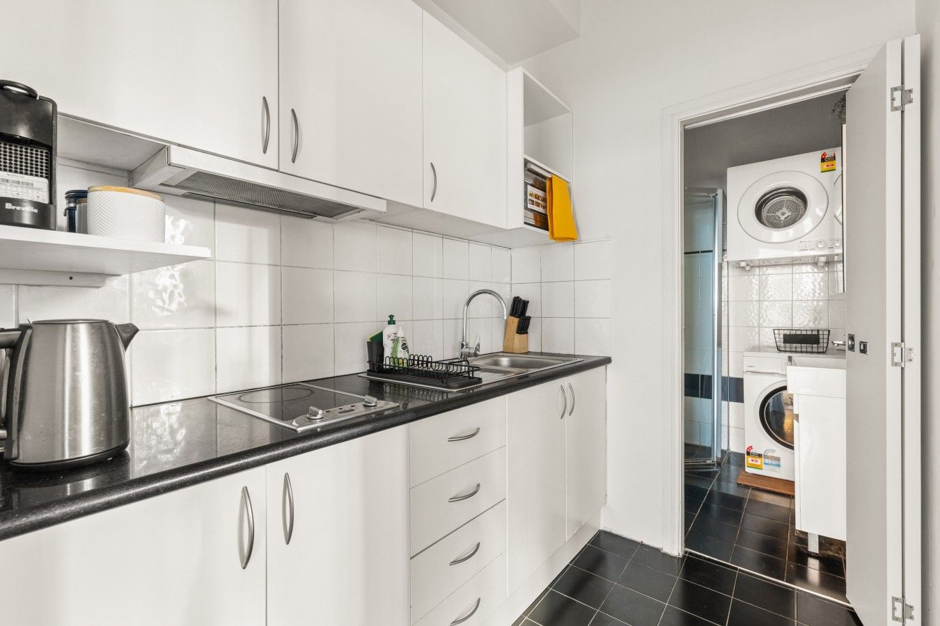 A modern kitchen with white cabinets, a black countertop, a kettle, and a washing machine in the background.