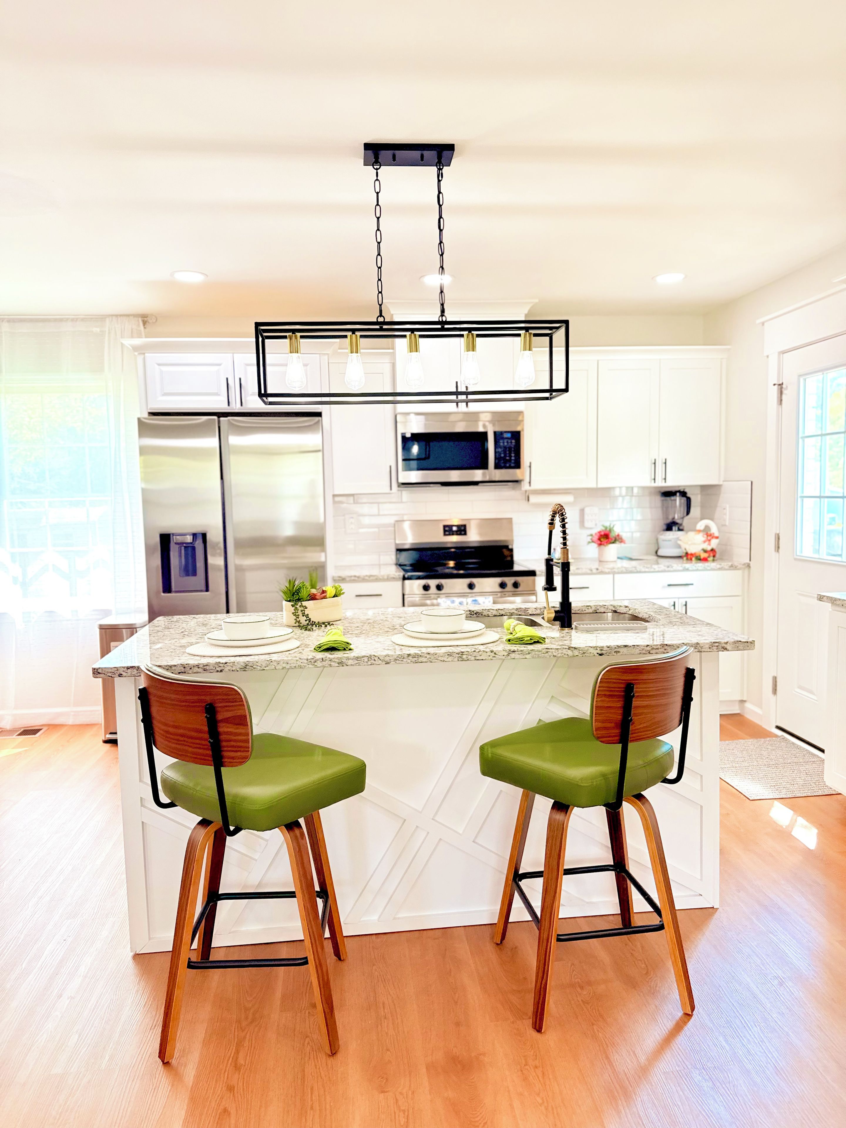 A modern kitchen with white cabinetry, stainless steel appliances, a granite countertop, and two bar stools with green cushions.