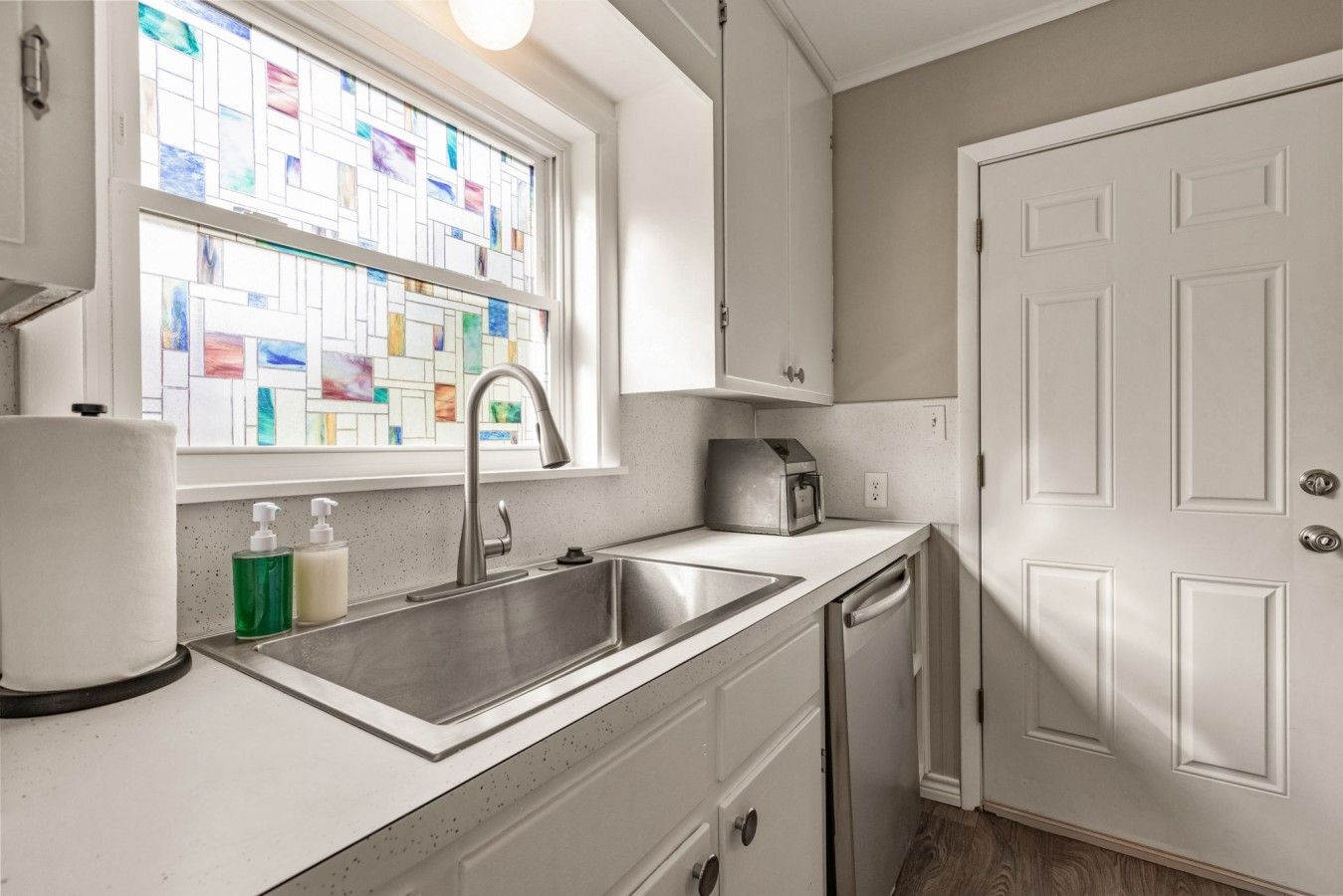 A kitchen with a stainless steel sink, a window with colorful stained glass, and a white door.