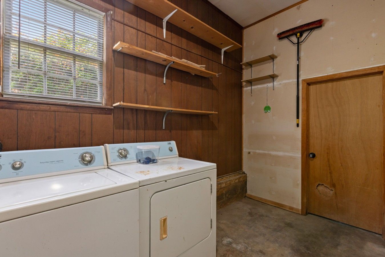 A laundry room with a washer and dryer, wooden shelving, a broom, and a door.