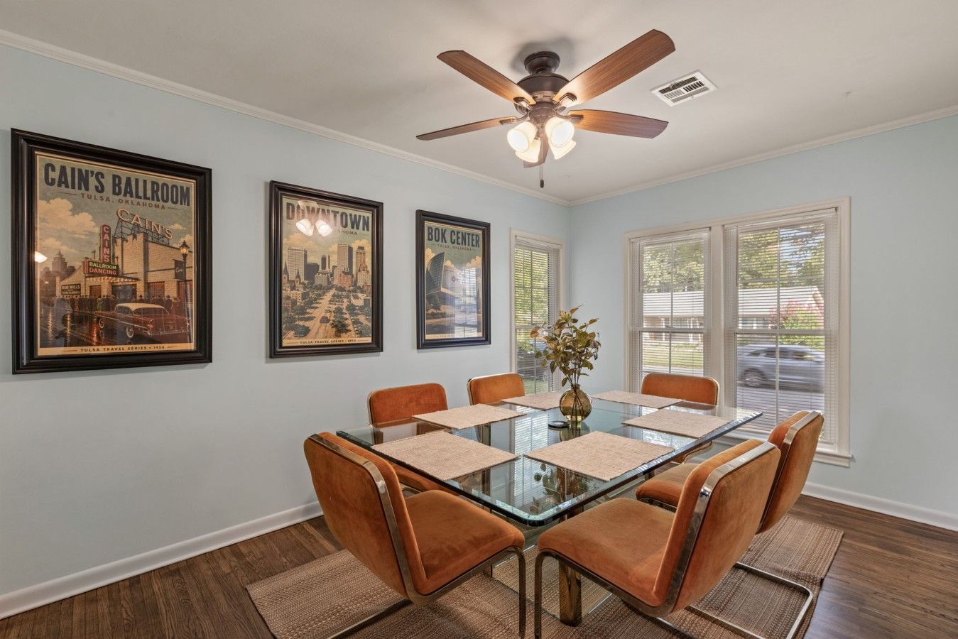 A dining area with a glass-top table, six chairs, a ceiling fan, and framed cityscape posters on the wall.