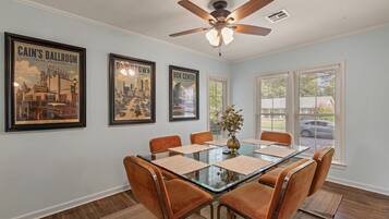 A dining area with a glass-top table, six chairs, a ceiling fan, and framed cityscape posters on the wall.