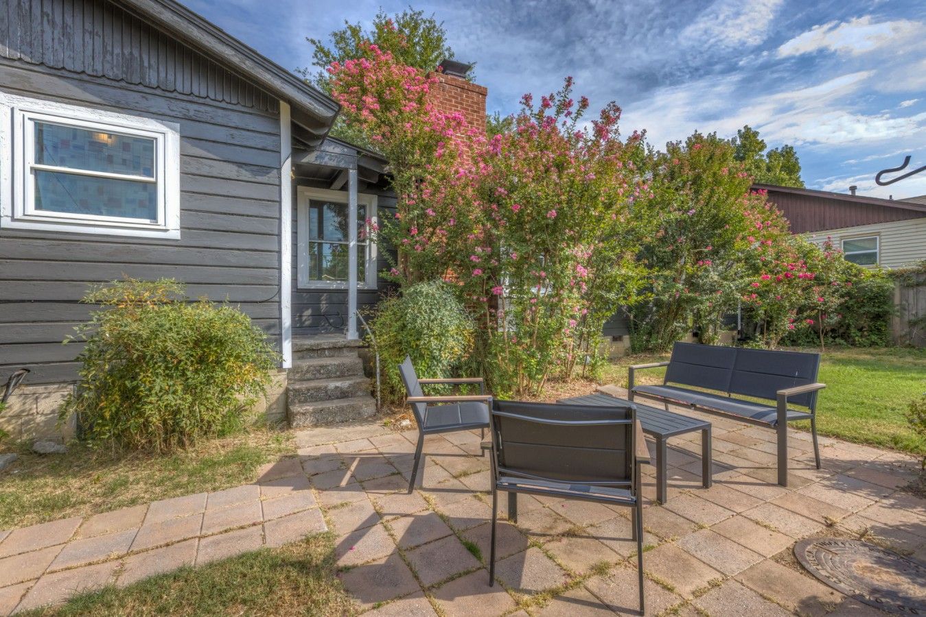 A patio with a bench and a small table, a wooden house with a porch, and a flowering bush.