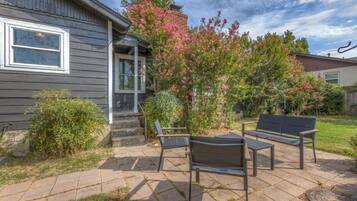 A patio with a bench and a small table, a wooden house with a porch, and a flowering bush.