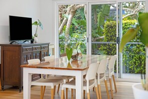 A dining area with a wooden table and white chairs, a television on a cabinet, and a view of lush greenery through large glass doors. - The Blue House Spacious Home (Port Macquarie)