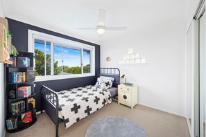 A bedroom with a bed, bedside table, bookshelves, and a window with a view. - The Blue House Spacious Home (Port Macquarie)