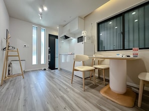 Ein modernes Hotelzimmer mit Holzboden, einem Essbereich mit zwei Stühlen, einem kleinen Tisch, einem an der Wand montierten Fernseher und einem fenster mit Vorhängen.