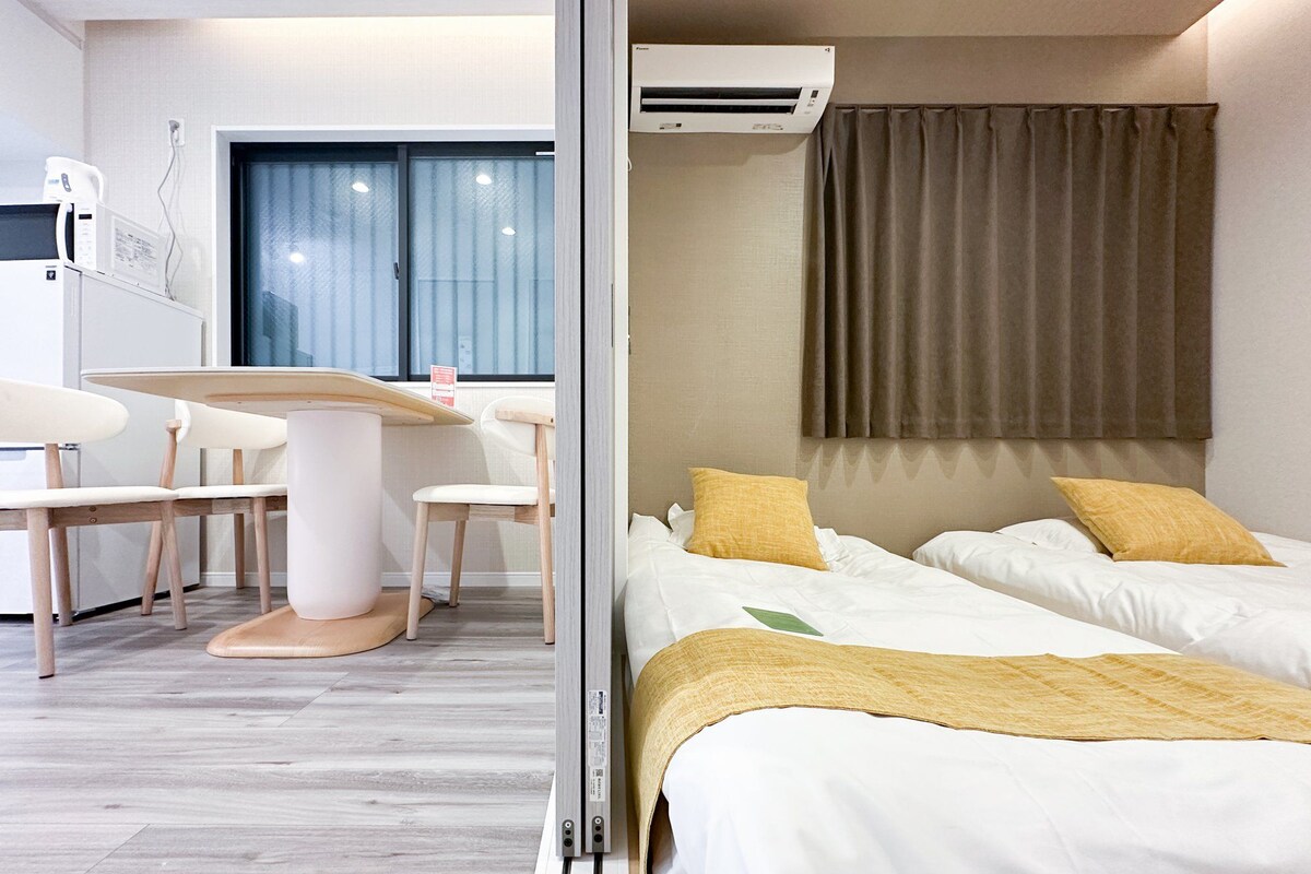Ein Hotelzimmer mit einem Bett, einem Schreibtisch und einem Stuhl. Ein Fenster mit Vorhängen und eine Klimaanlage befinden sich über dem Bett.