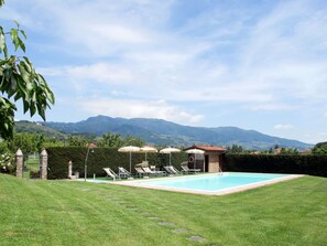 Una piscina con sillones de jardín y sombrillas, rodeada de un césped cuidado y un muro de piedra.