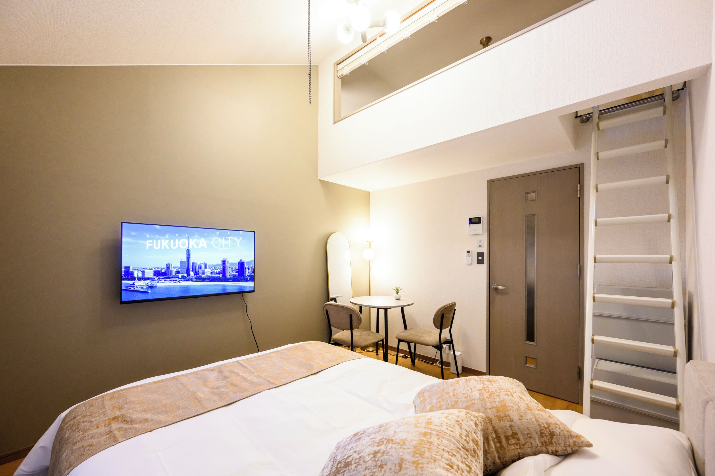 A hotel room with a bed, a television displaying a cityscape, a small table with chairs, and a door with a towel rack.