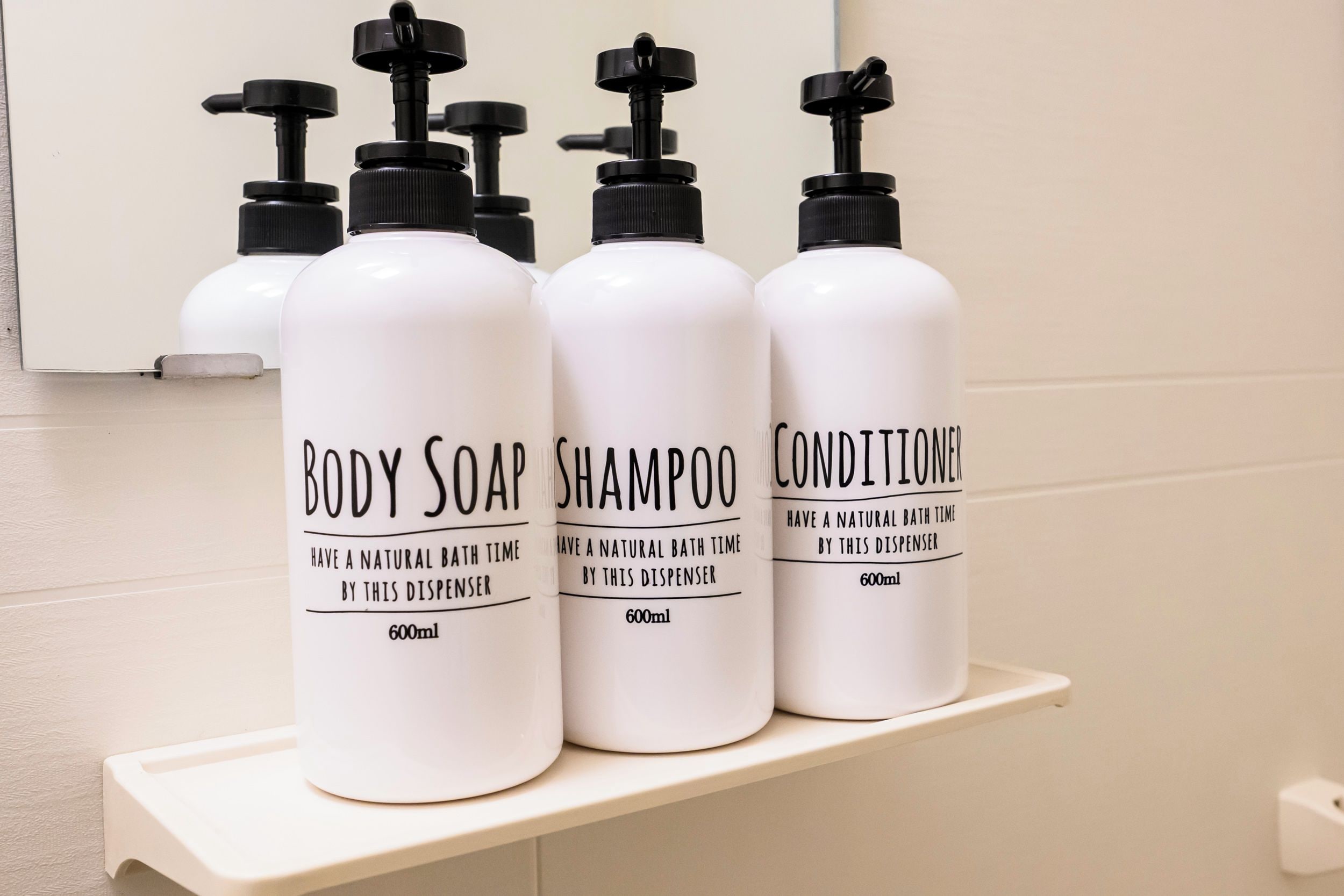 Kolme valkoista pulloa, joissa on vartalosaippuaa, shampoota ja hoitoainetta, valkoisella hyllyllä.