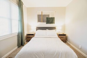 A bedroom with a large bed, two bedside tables, a wall-mounted light fixture, and a window with curtains. - Spacious Meridian Hills Retreat | Quiet & Private (Meridian Hills)