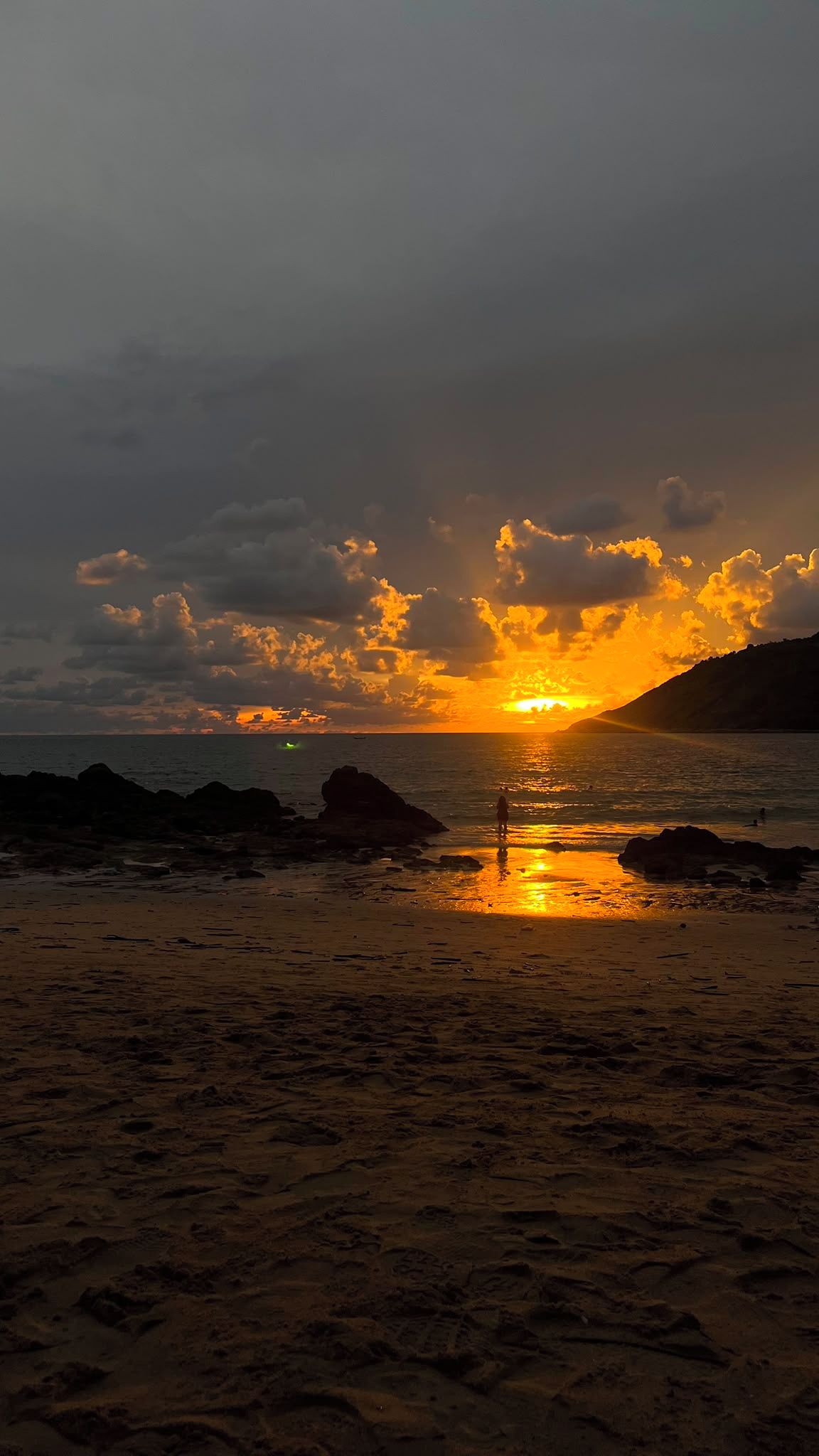 Een zonsondergang boven het strand, met een rotsachtige kustlijn en een persoon die in het water staat.