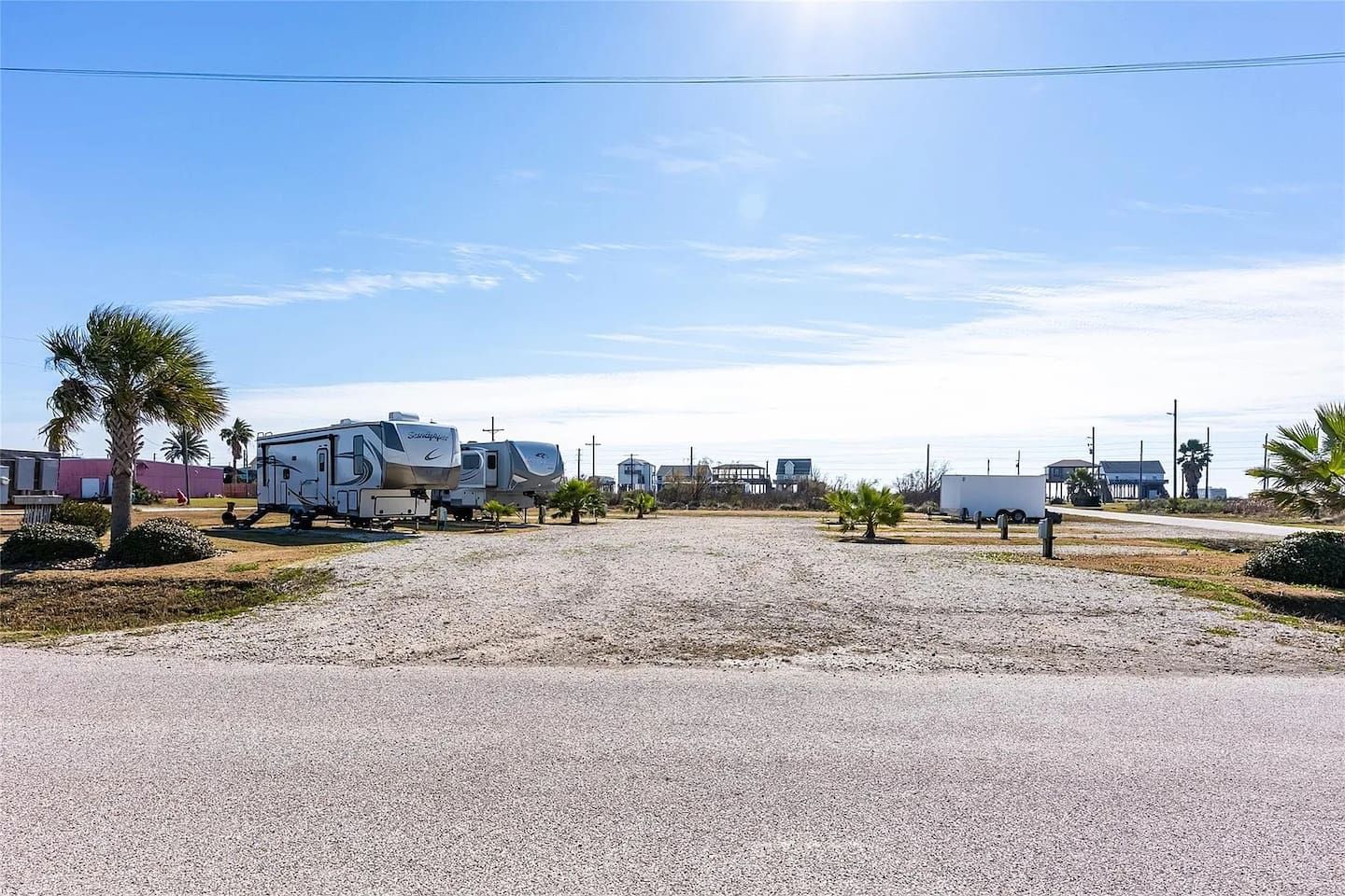 A gravel parking area with RVs and trailers, a clear sky, and sparse vegetation.