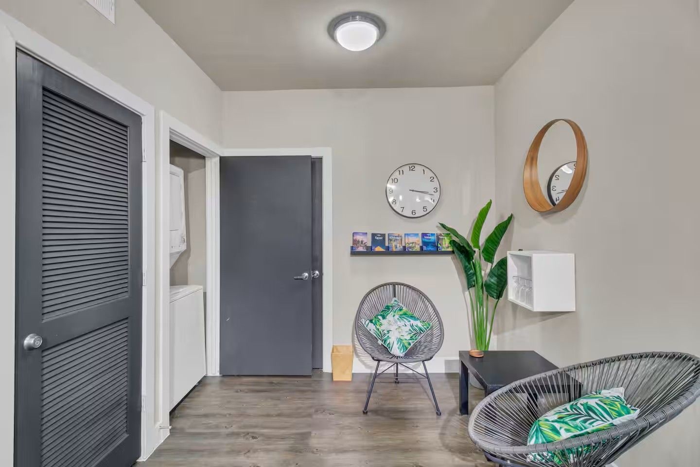 A hallway with a closed door, a wall-mounted clock, a shelf with books, a potted plant, and a wicker chair.