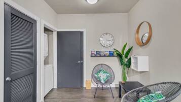 A hallway with a closed door, a wall-mounted clock, a shelf with books, a potted plant, and a wicker chair.