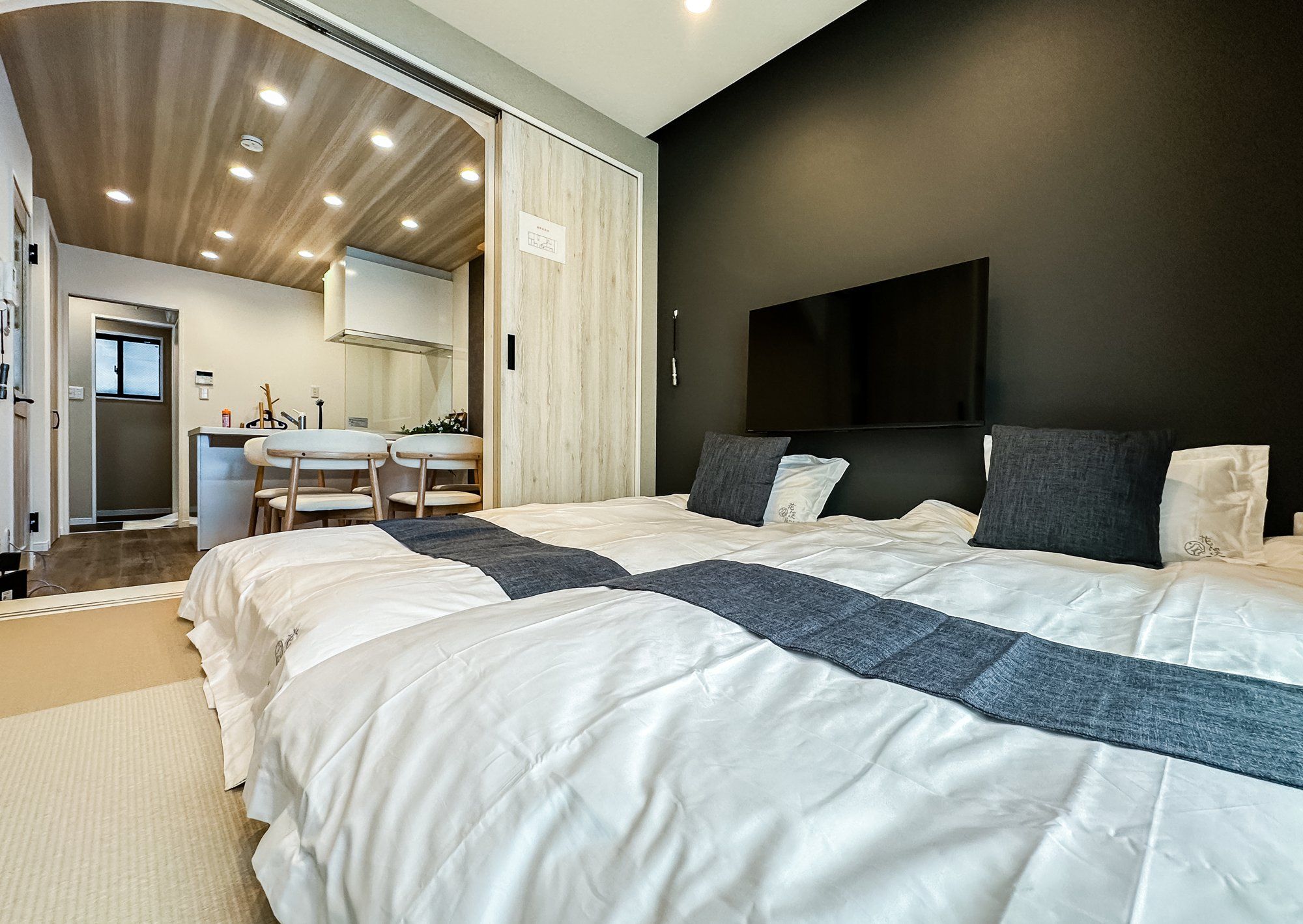 A modern bedroom with a large bed, a flat-screen TV mounted on the wall, and a kitchenette area visible through an open door.