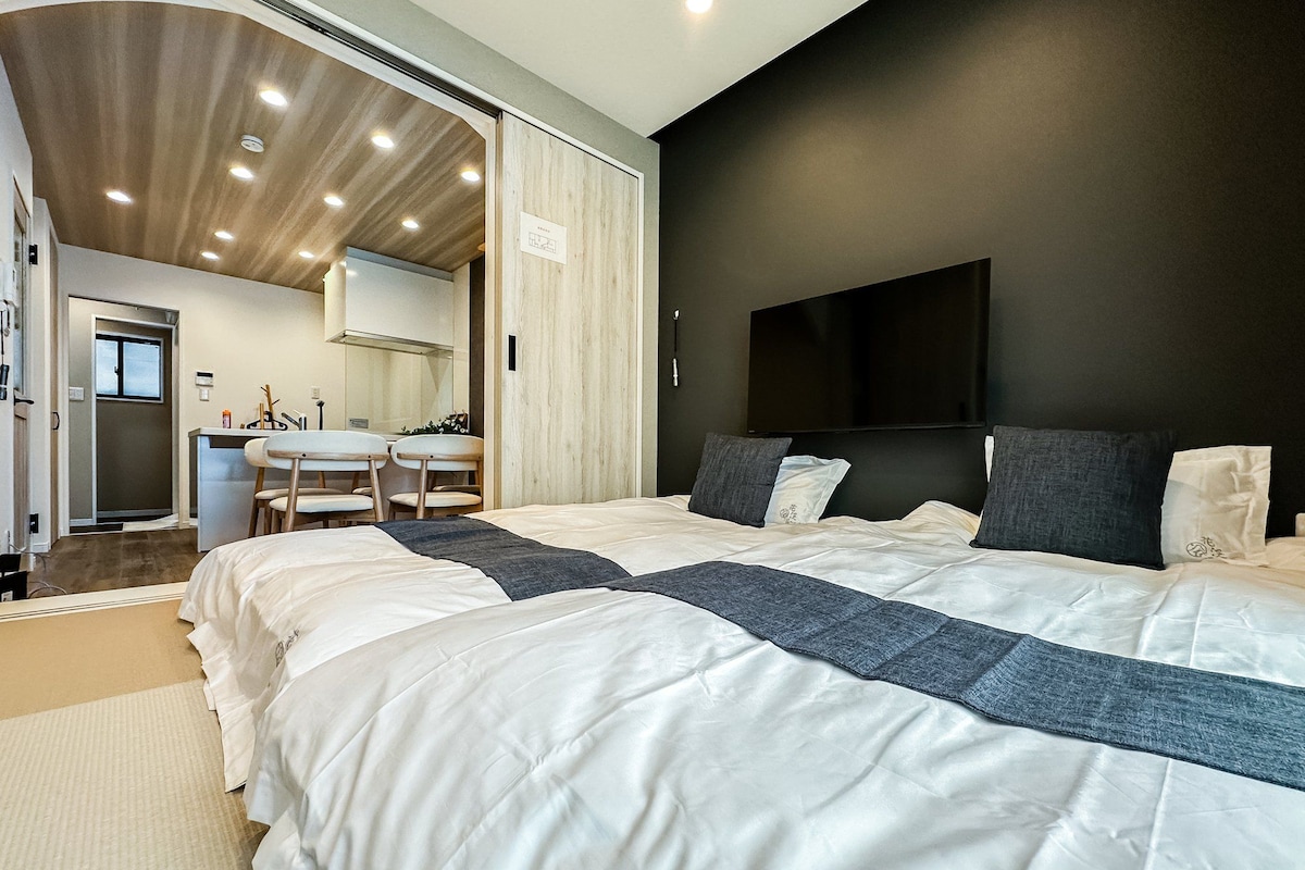 Ein modernes Schlafzimmer mit einem großen Bett, einem flachbildfernseher an der Wand und einer sichtbaren Küchenzeile durch eine offene Tür.
