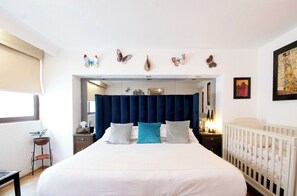 A bedroom with a bed, a crib, a nightstand, a window with a blind, and butterfly wall art. - Polanco Connected | South, North & Mexico City (Mexico City)